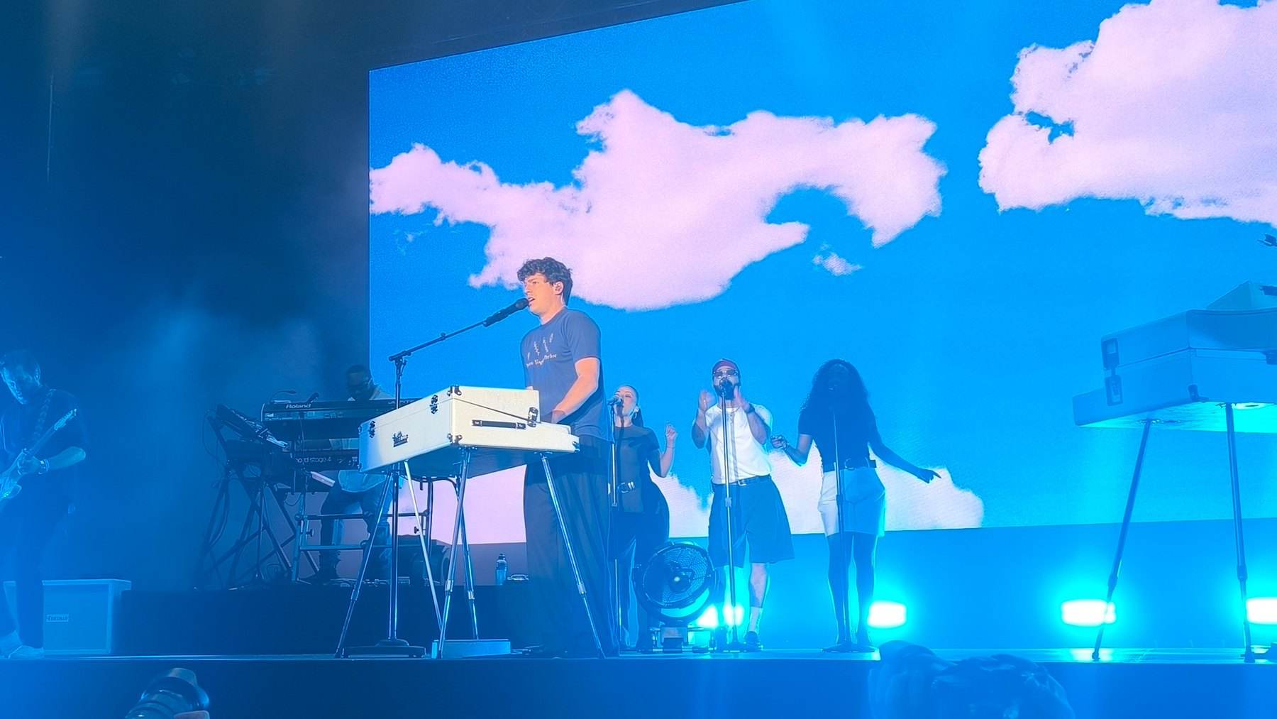 Charlie Puth singing at a keyboard on stage with his band, in front of a blue-sky screen